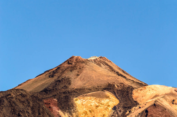 Kráter nejvyššího vrcholu za formacemi lávových skal na slunném a velmi jasném dni v národním parku El Teide. 13. dubna2019. Santa Cruz De Tenerife Španělsko Afrika. Cestovní ruch Street fotografie. - Fotografie, Obrázek