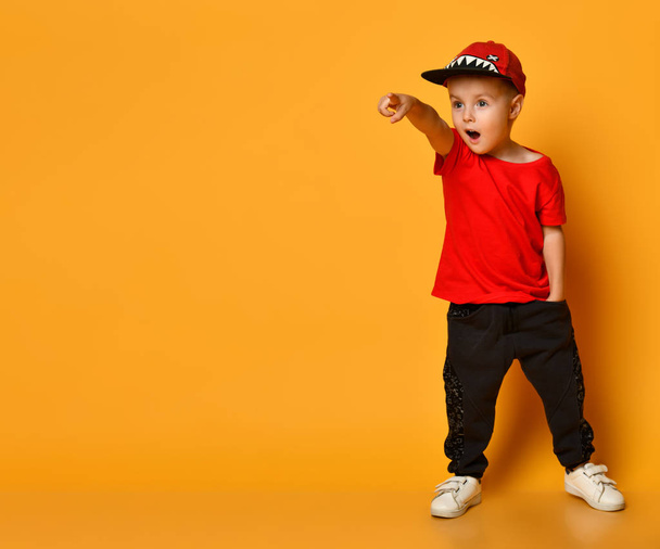 cute 4 year old boy in wide dark trousers with pockets and a red T-shirt. Funny hat with shark teeth. Standing on a yellow background shows a finger at something interesting. - Fotografie, Obrázek
