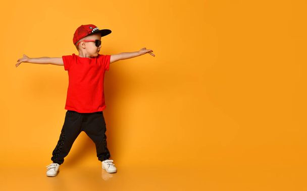 Young guy boy in a red T-shirt and dark pants, white sneakers and a funny cap posing on a free copy space on a yellow background - Fotografie, Obrázek