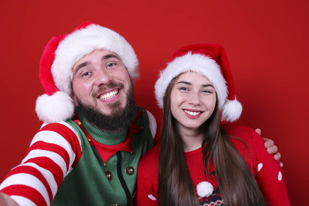 Smiling young couple wearing santa hats on red wall background. - Фото, зображення