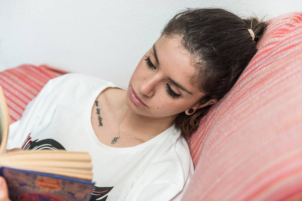 Tmavovlasá dívka tiše čte knihu sedící na gauči. - Fotografie, Obrázek