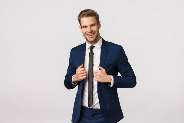 Confidence, elegance and business concept. Handsome young stylish businessman in suit, adjusting jacket and smiling, signed great deal and feeling success coming his hands, white background - Photo, Image
