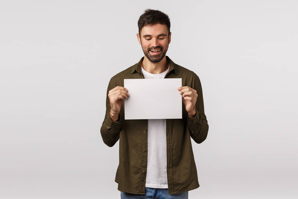 Satisfied, pleased charming handsome caucasian man with beard in modern outfit, have something to offer, holding blank piece paper, recommend visit web page, look at writing with pleased smile - Fotó, kép