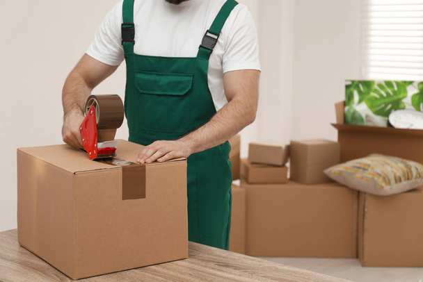 Man packing box with adhesive tape indoors, closeup. Moving serv - Фото, изображение