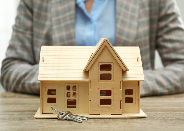 Real estate agent with house model and keys at wooden table, closeup - 写真・画像