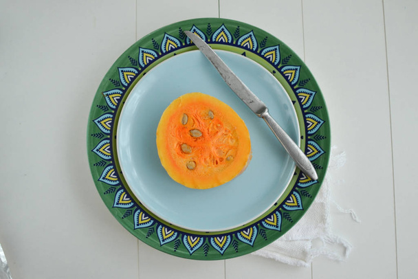Een schijfje pompoen met zaden op een turquoise bord met een metalen mes op een geruite servet - Foto, afbeelding