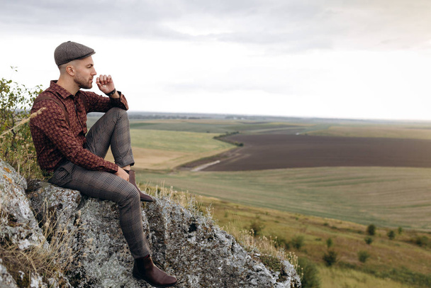 Εικόνα ενός νεαρού επιχειρηματία, αγρότη που κάθεται στην κορυφή του βράχου και ατενίζει την απόσταση από το όμορφο τοπίο των δικών του χωραφιών, σκεπτόμενος τα μελλοντικά σχέδια. - Φωτογραφία, εικόνα