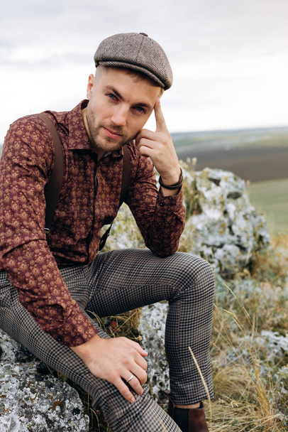 Portrait of pensive man in shirt, cap and suspenders, who sits on the rock top of the hill. Man is looking on camera. Beatuful landscape on background - Zdjęcie, obraz