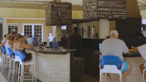 View on exterior of outdoor bar. Tourists on high blue white bar chairs around bar counter. Barman making bevereges and food. Oranjestad. Aruba. 09.14.2019 - Záběry, video