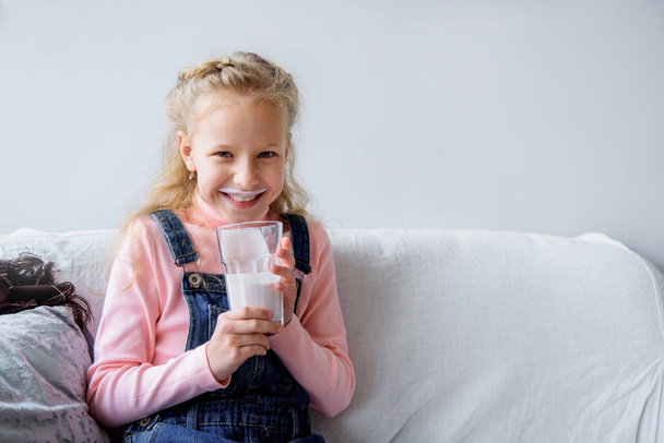 Girl drinks kefir or milk for breakfast. funny mustache. laughing on the couch at home. healthy eating children - Photo, Image