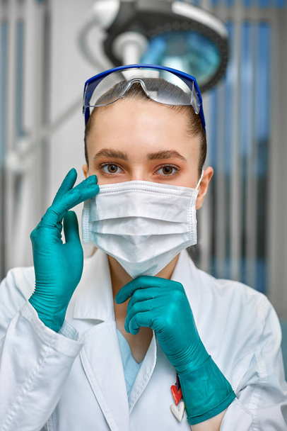 young beautiful woman doctor in white uniform and mask on her face close up on clinic background - Foto, Imagen