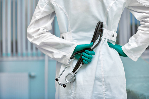 Doctor sosteniendo estetoscopio detrás de su espalda, retrato de cintura alta en el fondo de la clínica
 - Foto, imagen