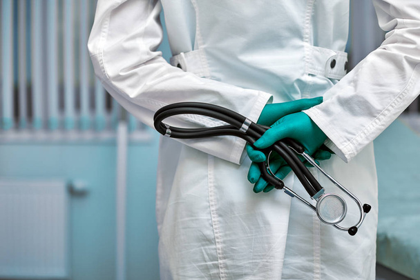 Arzt hält Stethoskop hinter dem Rücken, hüfthohes Porträt auf Klinikhintergrund - Foto, Bild