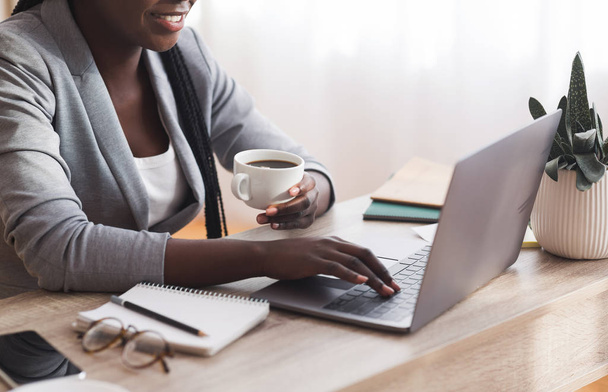 Empresária afro irreconhecível trabalhando no laptop e bebendo café no local de trabalho
 - Foto, Imagem