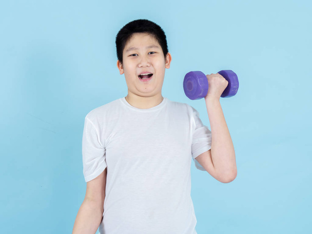 Smiling Asian boy using dumbbell on blue background. - Photo, image
