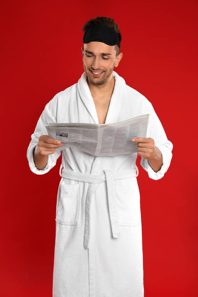 Happy young man in bathrobe reading newspaper on red background - Zdjęcie, obraz