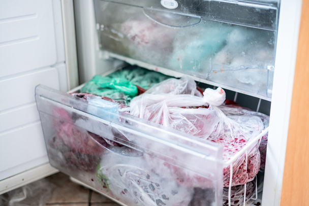 the opened freezer part of the fridge with many frozen packages of food - 写真・画像