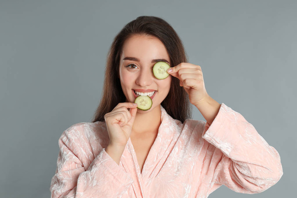 Young woman in bathrobe with cucumber slices on grey background - Фото, изображение