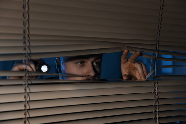 Male security guard with flashlight looking through window blinds in darkness - Фото, зображення