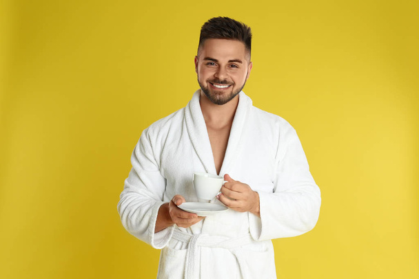Young man in bathrobe with cup of coffee on yellow background - Zdjęcie, obraz