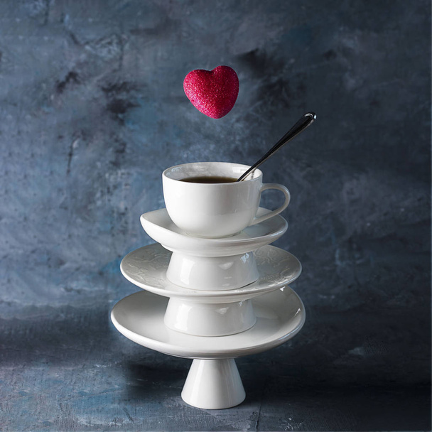 White saucer pyramid with cup of tea on top and flying red hearts candy over it. Dark background. Creative concept, love, relations, gift, celebrate, restaurant, food levitation, St. Valentine's day - Fotografie, Obrázek
