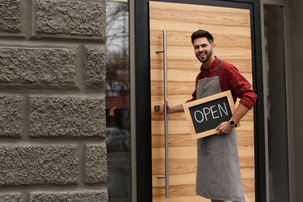 Young male business owner holding OPEN sign near his cafe. Space for text - Foto, immagini
