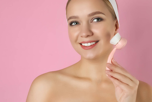 Young woman washing face with cleansing brush on pink background - Fotó, kép