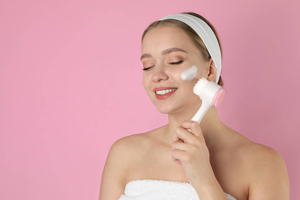 Young woman washing face with brush and cleansing foam on pink b - Photo, Image