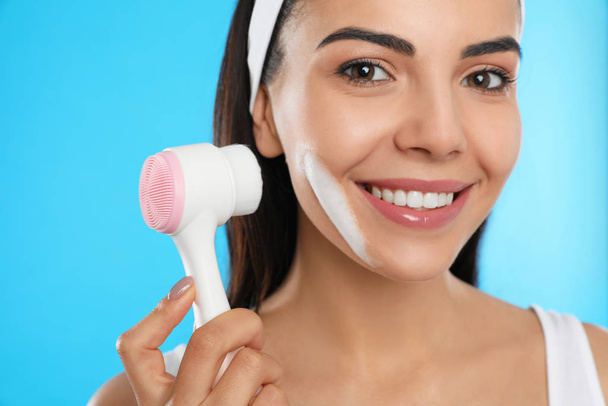 Young woman using facial cleansing brush on light blue background, closeup. Washing accessory - Foto, afbeelding
