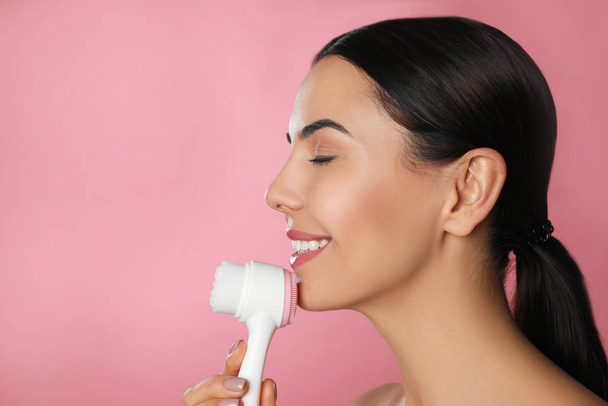 Young woman using facial cleansing brush on pink background, space for text. Washing accessory - 写真・画像