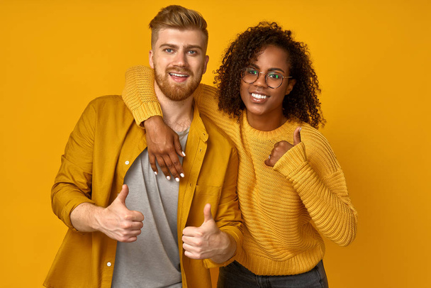 Horizontal shot of satisfying mixed race couple laughs happily, leans at the shoulder of boyfriend, come together on a theme party, dressed in yellow clothes. Pleased bearded Afro man shows thumb up - Foto, Bild
