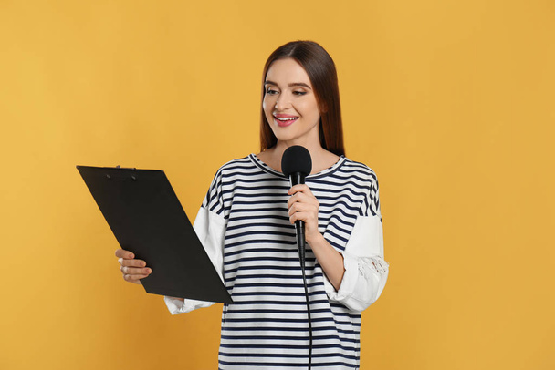 Young female journalist with microphone and clipboard on yellow background - Фото, изображение