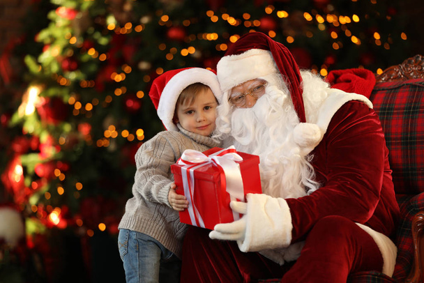 Santa Claus and little boy with gift near Christmas tree indoors - Photo, Image