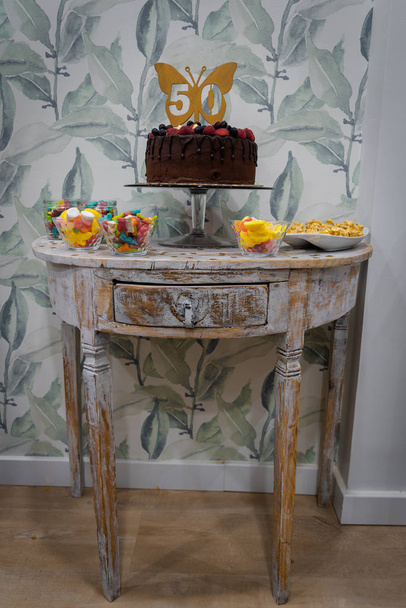 50の誕生日のチョコレートケーキは、驚き、お祝い、グミ、サーペンタイン、コンフェッティなどのパーティーのために装飾されたヴィンテージテーブルに提示されます.. - 写真・画像