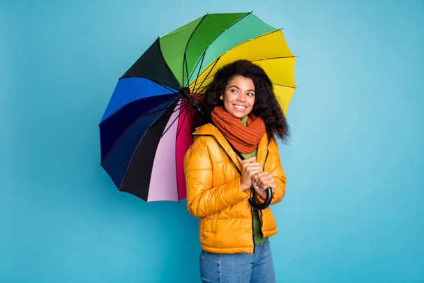 Photo of beautiful dark skin wavy lady good winter weather walk street hold umbrella look empty space wear yellow coat jeans sweater scarf isolated blue color background - Foto, imagen