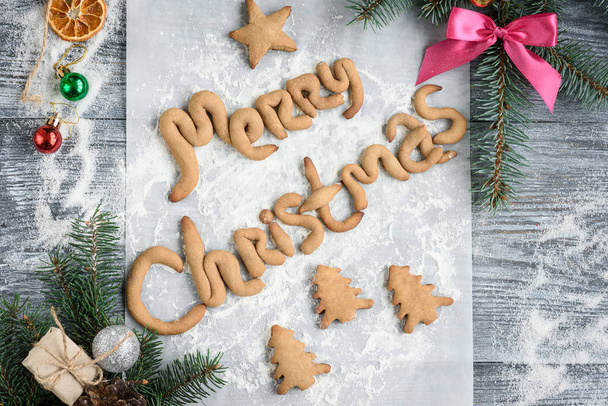 Vrolijke kersttekst van zelfgebakken peperkoek. Kerst samenstelling van sparren takken, kegel, boog, ballen en peperkoek koekjes op grijze houten plank. - Foto, afbeelding