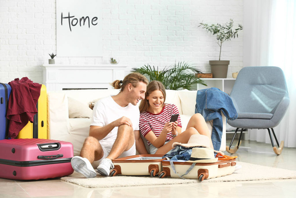 Young couple with mobile phone packing suitcases at home - 写真・画像