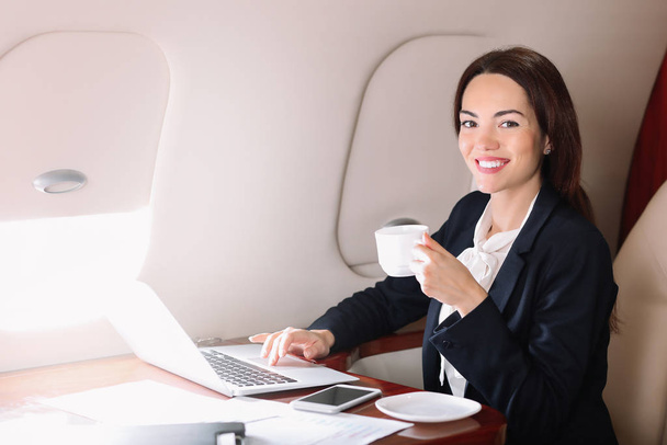 Zakenvrouw met laptop die koffie drinkt aan boord van het moderne privévliegtuig - Foto, afbeelding