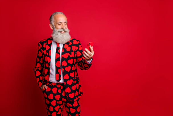 Photo of funny mature guy holding telephone chatting girlfriend write love sms letter wear stylish hearts pattern suit blazer shirt tie pants isolated red color background - Foto, Imagem