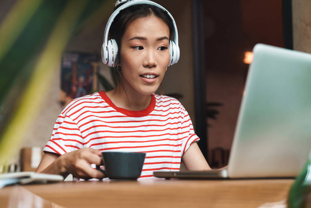 Portrait of satisfied asian woman with headphones using laptop i - Zdjęcie, obraz