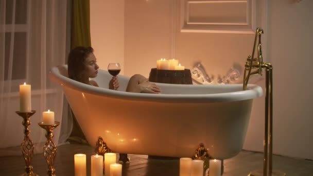 Женщина лежит в пенной ванне со свечами, чтобы расслабиться и выпить вина. Романтическая расслабляющая атмосфера ароматерапии. Ванная комната со свечами
 - Кадры, видео