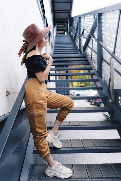 Μοντέρνα μελαχρινή γυναίκα με μακριά μαλλιά, φοράει κομψό μεγάλο ψάθινο καπέλο, ποζάροντας στις σκάλες - Φωτογραφία, εικόνα