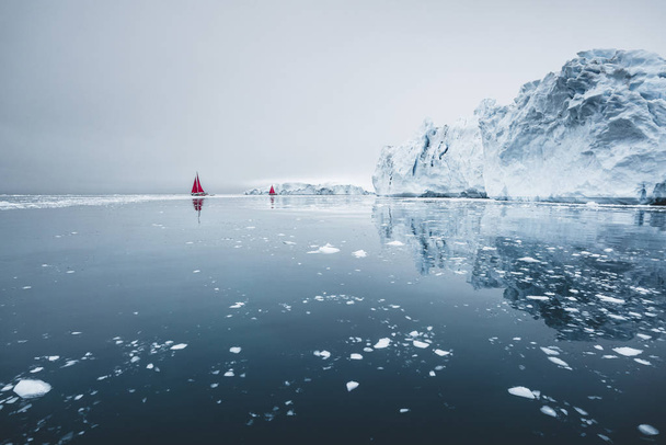 Beautiful red sailboat in the arctic next to a massive iceberg showing the scale. Cruising among floating icebergs in Disko Bay glacier during midnight sun season of polar summer Ilulissat, Disko Bay - 写真・画像