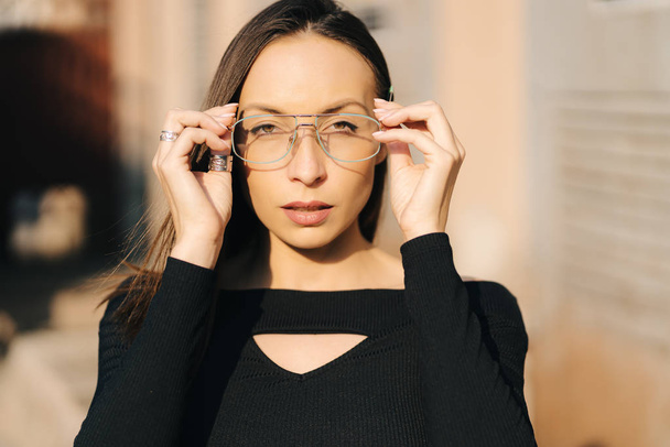 Atrakcyjna młoda brunetka w czarnej bluzce i przezroczystych okularach pozujących na ulicy - Zdjęcie, obraz