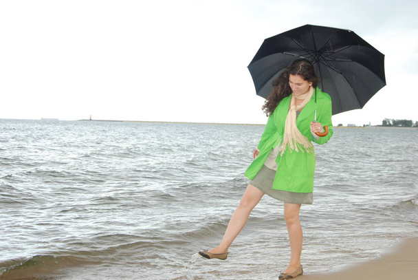 The girl in a bright green jacket with a black umbrella at the beach on the horizon. Cold weather at the sea, storm. - Foto, Imagem