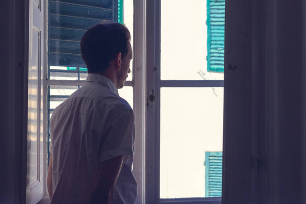 Mann blickt aus Fenster eines dunklen Zimmers  - Foto, Bild