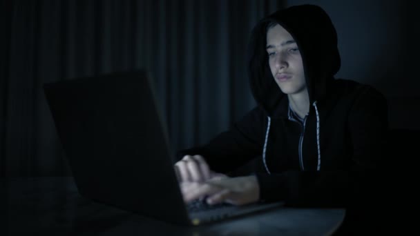 Hacker's hacking into the computer network. A young guy in a dark room typing on a laptop.  Hacker with a laptop. E-the robber. Lonely teen chatting online. Computer criminal. 4K footage - Metraje, vídeo