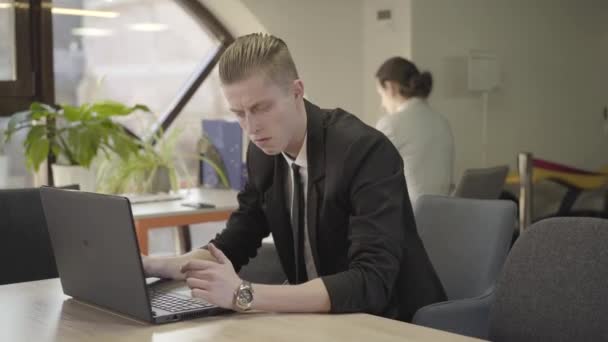 Portrait of young Caucasian man sitting in open space office and typing on laptop keyboard. Stressed male office worker dealing with business issues at workplace. Business, CEO, management. - Séquence, vidéo