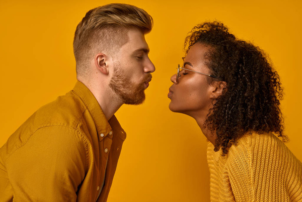 Mixed race couple stand in profile, keep lips folded, staring at each other, going to kiss, dressed casual mustard color wear, make grimace, pose over orange wall, have fun. Facial expressions - Foto, immagini