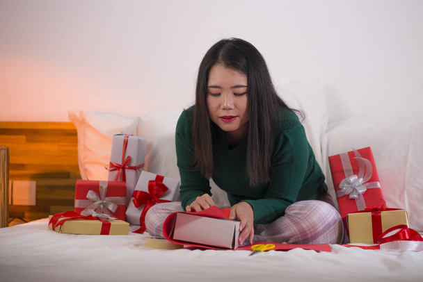 Christmas holiday lifestyle portrait of young happy and sweet Asian American woman sitting on bed preparing xmas presents wrapping gift boxes and adding ribbons - Photo, image